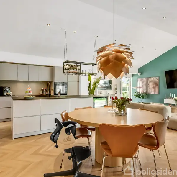 Rundt spisebord med moderne stole står i et åbent køkken-alrum. En stor kobberfarvet loftslampe hænger over bordet. Et køkken med ø og sofaområde ses i baggrunden.