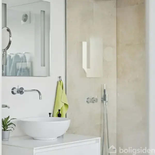 Et hvidt badeværelse viser en rund vask med en vægmonteret vandhane. Ved siden af hænger et grønt håndklæde. I baggrunden er en glasbruseniche.