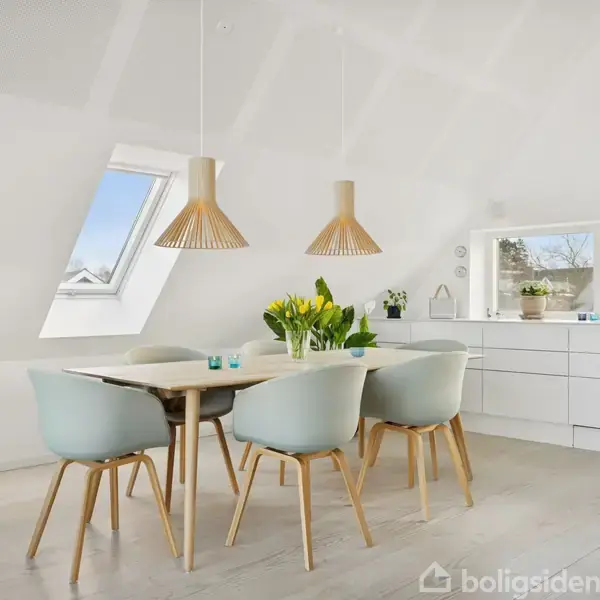 Et spisebord i lyst træ omgivet af fire blå stole står under to trælamper. Bagved ses et vindue og en hvid skænk med planter, skabende en lys og moderne indretning.