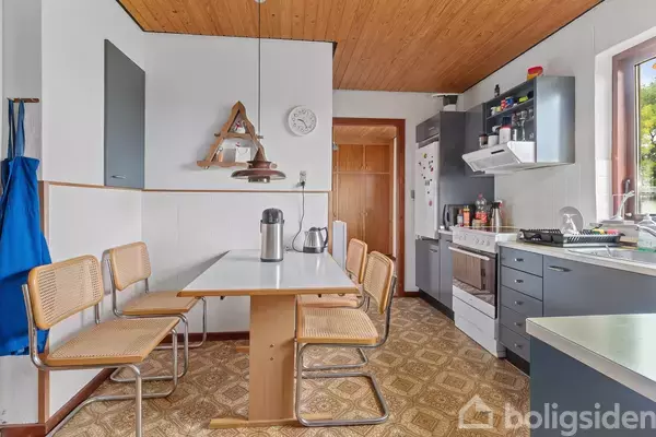 Et køkken med et rektangulært bord omgivet af fire stole. Bordet har en termokande i midten. Køkkenet har grå skabe, en ovn og en håndvask ved vinduet.