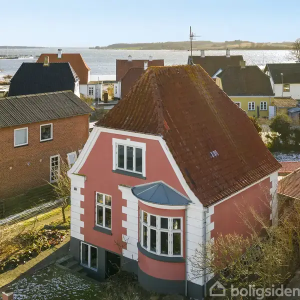 Et lyserødt hus med rødt tag står blandt andre huse i et kystområde. Ved siden af huset er en parkeret bil. Skiltet på en bygning viser teksten "GRILL" i baggrunden.