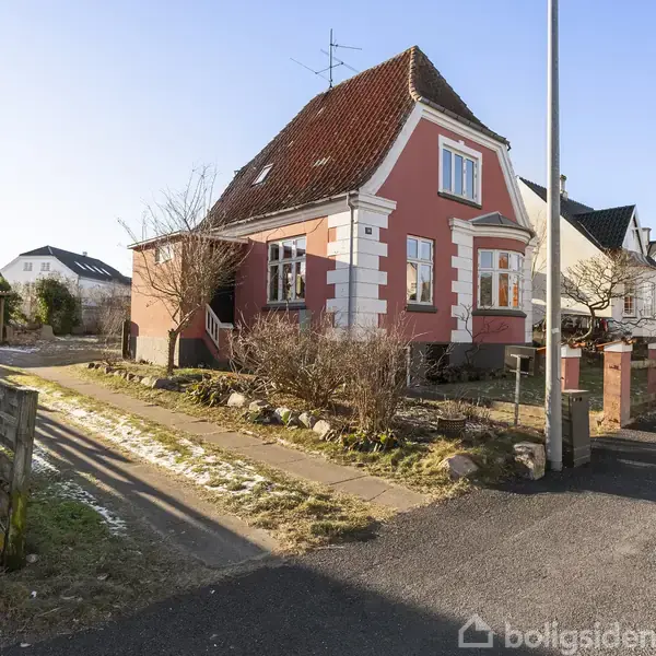 Et lyserødt hus med brune teglstenstag står i et boligkvarter. Huset er omgivet af en have med buske og en gangsti langs vejen.