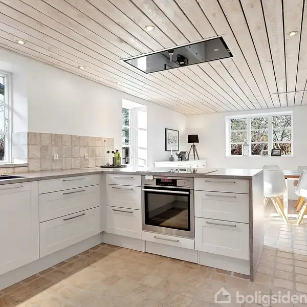 Et lyst køkken med hvide skabe og ovn indbygget i et L-formet skab. Rummet har træloft og flisegulv, store vinduer og åbner mod en spisestue med hvide stole og et bord.