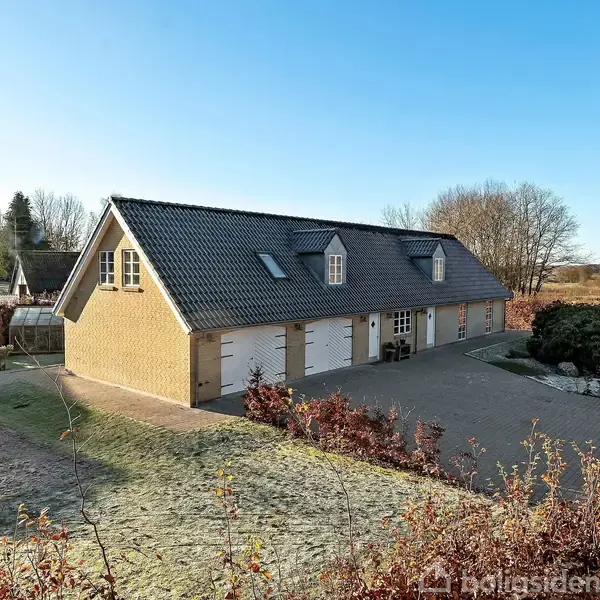 Et gult murstenshus med sort tegltag står i et åbent, frodigt landskab. Huset har vinduer og tre garagedøre, omkranset af buske og træer på en solrig dag.