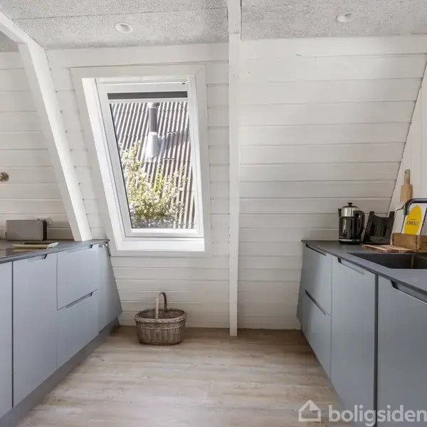 Gråt køkken med moderne skabe, arrangeret i en vinkel mod et skråtloftet vindue. Lys træpanelvægge og gulv. Køkkenredskaber og en kaffemaskine ses på den ene bordplade.