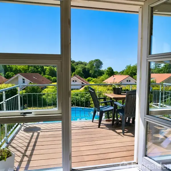 Balkon med bord og stole åbner ud mod en udsigt over grønne træer og rødt tageklædte huse i solskinnet, synligt fra et åbent vindue.