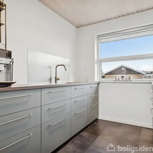 Et moderne køkken med lysegrå skabe og en indbygget kaffemaskine. Håndvasken er placeret ved et vindue, og rummet har hvide vægge og et lyst træloft.