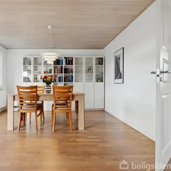 Træbord med seks stole er placeret i et lyst rum. Vaser med blomster står på bordet, og en bogreol fyldt med bøger står i baggrunden. Dørskiltet viser "HOME".