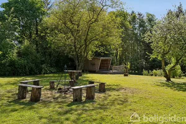 Bænke lavet af træstammer er arrangeret rundt om en bålplads i en solrig have. I baggrunden ses en træhytte omgivet af frodig vegetation.