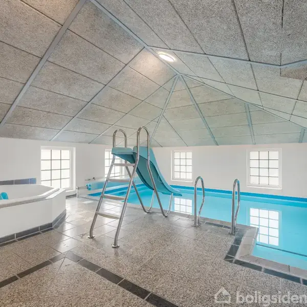 Swimmingpool med tilhørende rutsjebane og spabad i et indendørs rum med højt til loftet og store vinduer, der giver naturligt lys. Lyst klinkegulv og moderne interiør kompletterer omgivelserne.