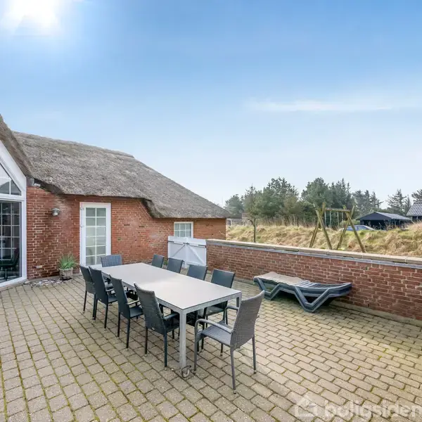 En terrasse med et langt spisebord og otte stole står på brosten. En murstensbygning med stråtag er baggrund, omgivet af natur med træer og huse i det fjerne.
