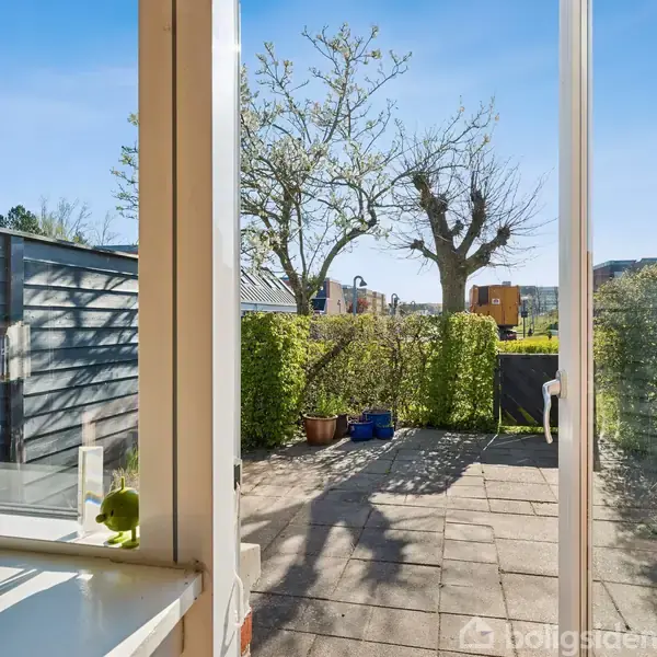 Et flisebelagt gårdhaveområde med et bord ses gennem en glasdør. I baggrunden er en træhæk og et stort, bladløst træ. Klar blå himmel og bygninger er synlige i horisonten.