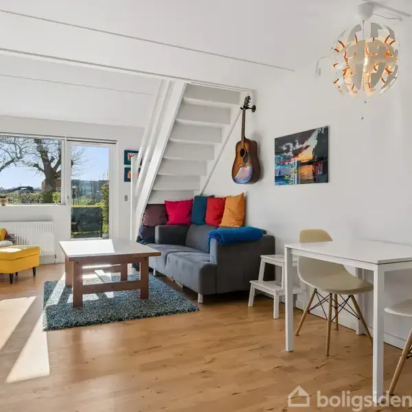 Stue med gråt sofaarrangement fyldt med farverige puder, en guitar hængende på væggen, gul lænestol ved stort vindue, spisebord med to stole, alt på trægulv.