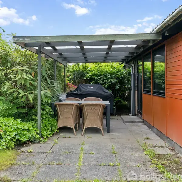 Terrassemøbler står under en overdækket pergola, omgivet af frodig beplantning ved siden af en orange facade med store vinduer.