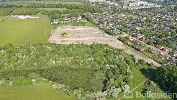 Luftfoto af et grønt område med skov og en åben mark, hvor et byggepladsområde ses centralt, omgivet af en bebyggelse af huse til højre og yderligere grønne arealer til venstre.