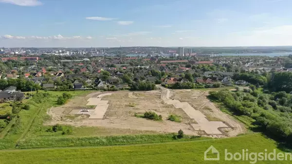 Byggegrund klar til udvikling, beliggende i et grønt område med udsigt over en by. Området er omgivet af træer og enkelte huse, med byen i baggrunden.