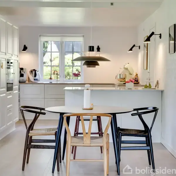 Køkken med et bord omgivet af fire stole. En stor lampe hænger over bordet. Køkkenet har hvide skabe, en vase med blomster og køkkenudstyr. Vinduet viser træer udenfor.