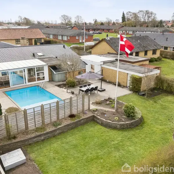 En villa med swimmingpool i haven, dansk flag på masten, omgivet af græsplæne og terrasse med møbler. Baggård understøtter et roligt boligområde med flere huse i baggrunden.