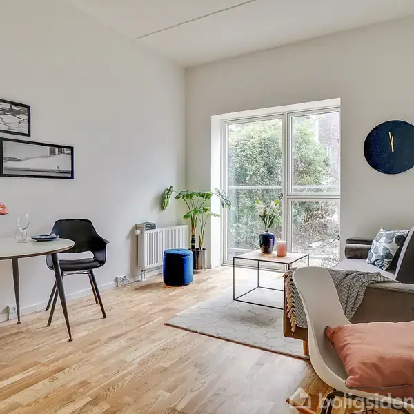 En stue med trægulv har en grå sofa og sofabord ved et stort vindue. Et rundt spisebord med blomster og vinglas står ved siden af. Væggene er dekoreret med kunst.