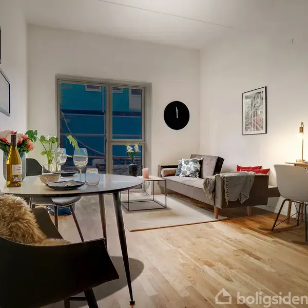 Et lille, moderne stueområde med trægulv. Et rundt bord står med glas og tallerkener, en sofa og skrivebord findes på den bagerste væg, omgivet af kunstplakater.