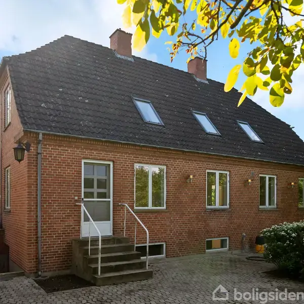 Rødt murstenshus med sort tag har vinduer og en hoveddør med trappe. Det står i et grønligt, solrigt område med flisebelagt gårdsplads og frodig bevoksning.