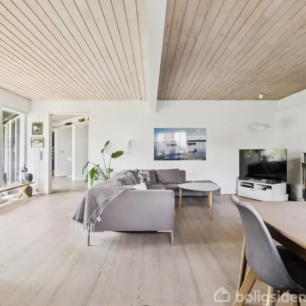 Stue med sofaer og spisebord ved siden af en pejs, omgivet af hvide vægge og store vinduer med udsigt til have. Lys træbeklædning på gulve og loft.