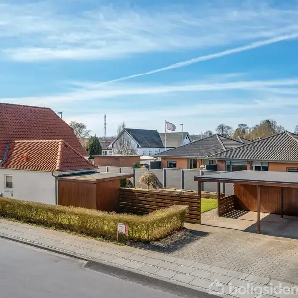 Et parcelhus med rødt tegltag står i forgrunden. En carport og en indkørsel er ved siden af. En hæk omkranser ejendommen i et beboelsesområde under en blå himmel.