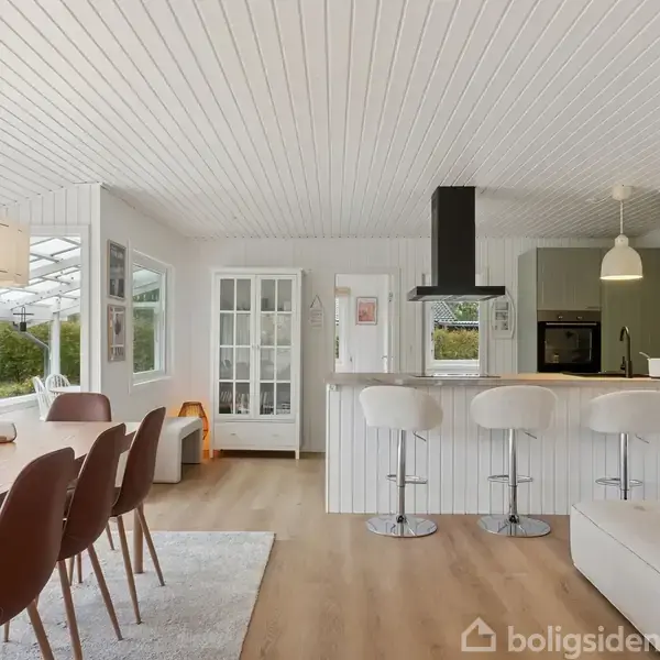 Et lyst køkken-alrum med hvidt træloft og trægulv. En spisebord med stole står ved siden af en sofa. Der er en køkkenø med barstole. Store vinduer giver udsigt til haven.