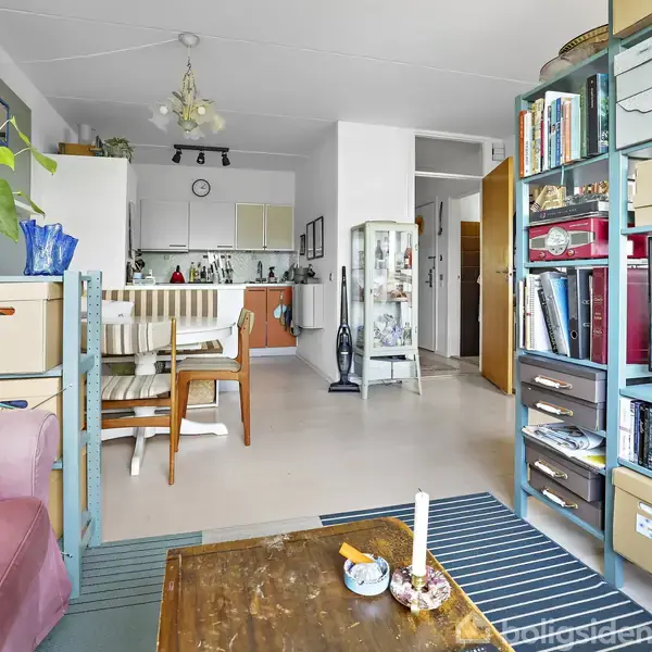Stue med reol fyldt med bøger og kasser, står ved siden af et fjernsyn. En lyserød sofa og et sofabord med lys står nær vindue, der giver belysning til rummet.