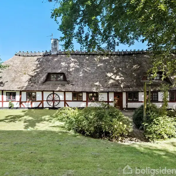 Bindingsværkshus med stråtag står i en solrig have omgivet af grønne træer og buske. En lille græsplæne breder sig foran huset, der har hvide vægge og brune træbjælker.