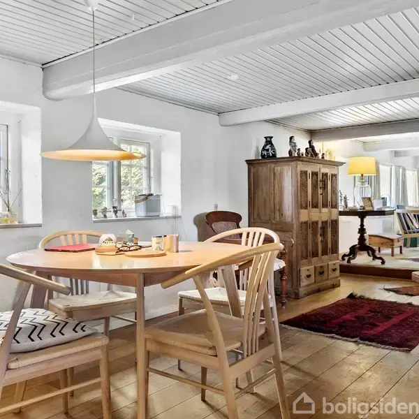 Et rundt spisebord med fire stole står på et trægulv. En pendellampe hænger over bordet. Vinduer giver naturligt lys, og et træskab og tæpper er i baggrunden.