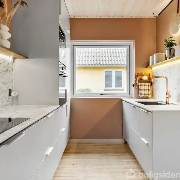 Et moderne, lyst køkken med hvide skabe og marmorbordplader. En indbygget kogeplade tilbereder mad. En stor vindue giver udsigt til et hus udenfor. Planter pynter rummet.