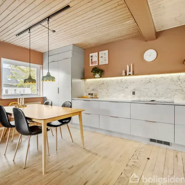 Træbord med sorte stole står i moderne køkken. Lyse skabslåger og marmorvæg skaber stilfuldt udseende. Træloftet og gulvet tilfører varme. Lamper hænger over bordet, og vindue viser træer udenfor.