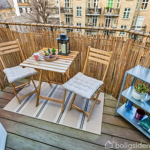 Balkon med to træstole og et bord, hvorpå der står en sort lanterne og en kop. En hylde til venstre med planter og lys. Omgivet af bambushegn i bymiljø.