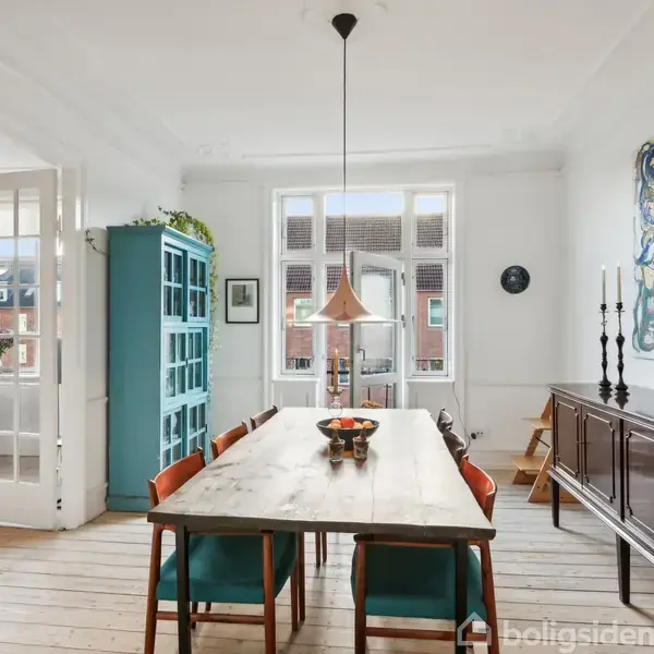 Et spisebord med stole står oplyst af en kobberfarvet lampe i et lyst rum med trægulv. I baggrunden ses et stort kunstværk på væggen og et blåt skab.