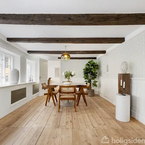 Et spisebord med stole står midt i et rum med trægulv og synlige loftsbjælker. Der er en pendellampe over bordet, planter og lysindfald fra vinduer til venstre.