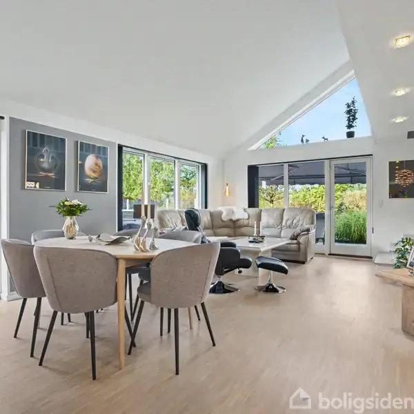 Stue med spisebord og stole, lejlighed med lædersofaer og moderne dekoration, store vinduer åbner mod en have, skaber en lys og indbydende atmosfære.