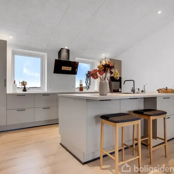 Et moderne køkken med en ø. To barstole står ved øen, pyntet med blomster og en vandhane. Skabe og vinduer omkranser rummet, der har lyst trægulv.