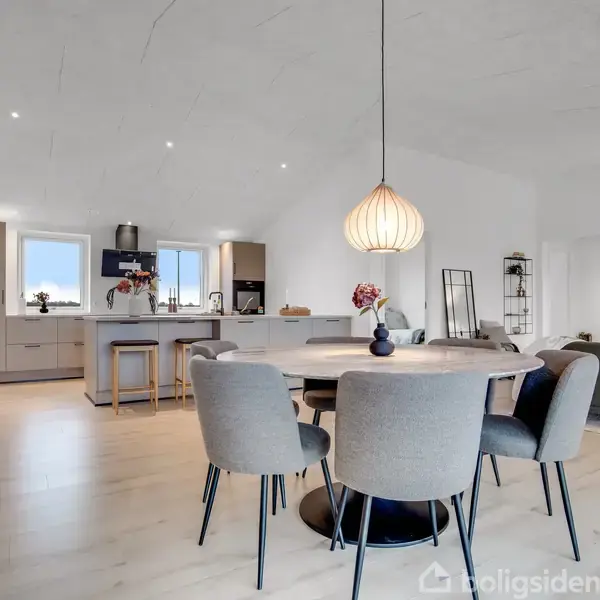 Spisebord med fire stole står centralt, mens en stor lampe hænger over det. Køkkenområdet med ø og hvide skabe ses i baggrunden. Rummet er lyst med moderne indretning.