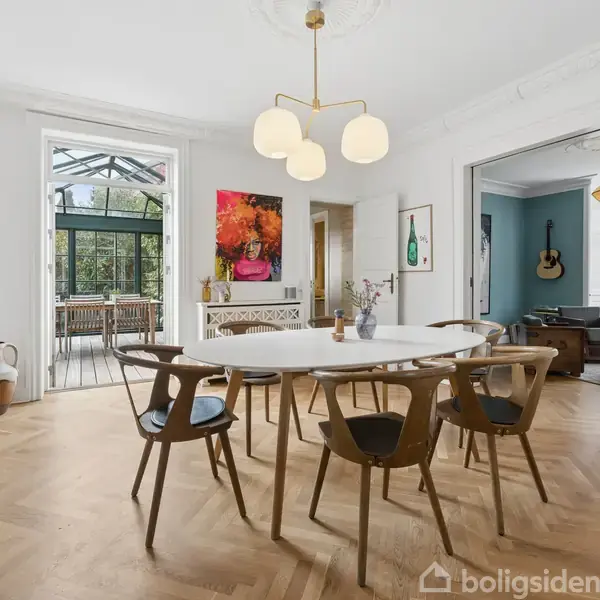 En rund hvid spisebord med seks stole står i en spisestue. En moderne lysekrone hænger over bordet. Rummet har trægulv og åbner ud til en terrasse med glasdøre.