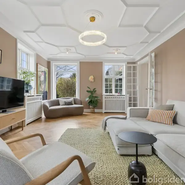 Stue med hvid sofa og rund bordlampe, fladskærms-TV på træmøbel, lænestol og runde sofabord. Store vinduer slipper lys ind, og væggen er dekoreret med kunstværker.