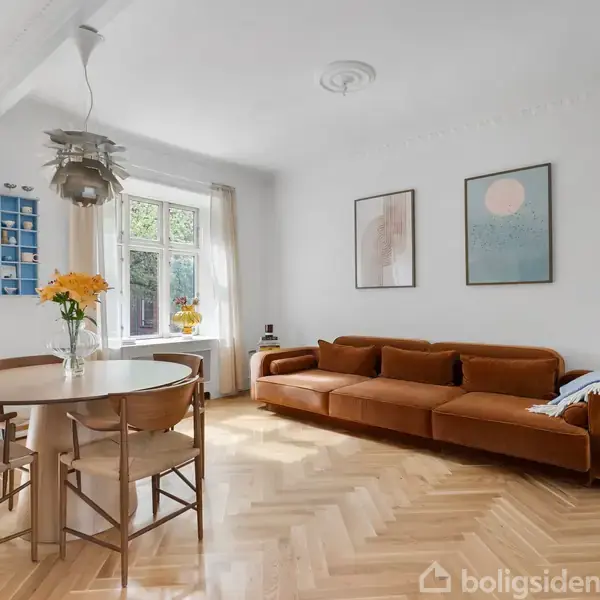 En brun sofa står mod en hvid væg med to abstrakte malerier. Ved siden af er et rundt spisebord med blomster i en vase, i et lyst rum med parketgulv.