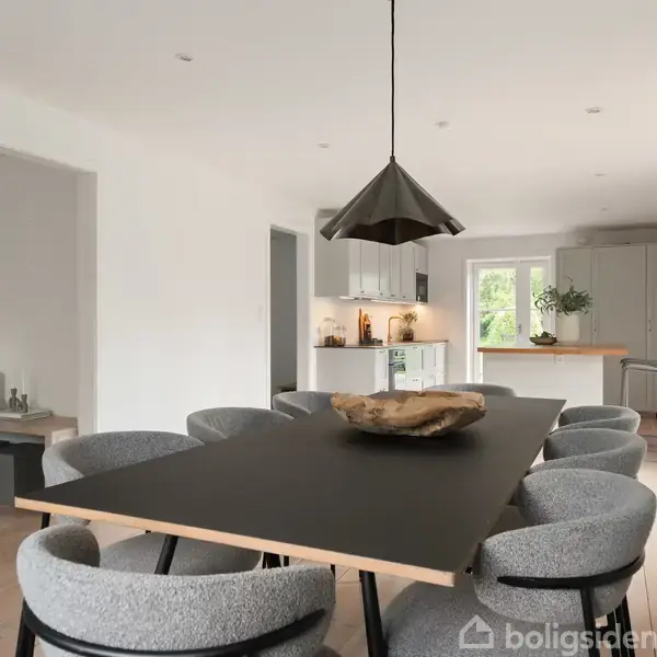 Et moderne spisestue med et stort, sort bord omgivet af seks grå stole. En lampe hænger over bordet. Rummet strækker sig til et åbent køkken i baggrunden med hvide skabe.