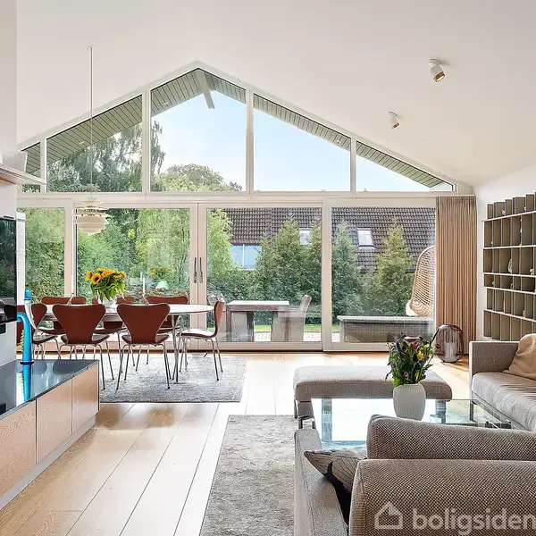 Stue med sofagruppe og glasbord er beliggende foran en stor vinduesvæg med udsigt til have. Ved siden af står en reol, spiseplads med stole og borddekorationer med blomster.