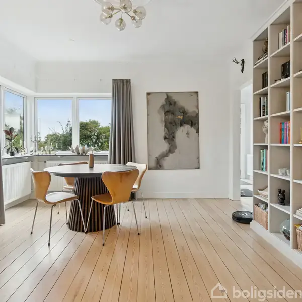 Rundt spisebord med fire stole står i et lyst rum med trægulv; hylder fyldt med bøger til højre og store vinduer bagved, der giver udsigt til træer udenfor.