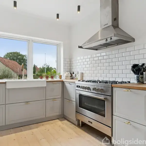 Et moderne køkken med grå skabe og flisevæg, indeholder en stor ovn og emhætte. Vinduet viser en udsigt til en hus og træer udenfor. Skærebræt og krydderier på bordet.