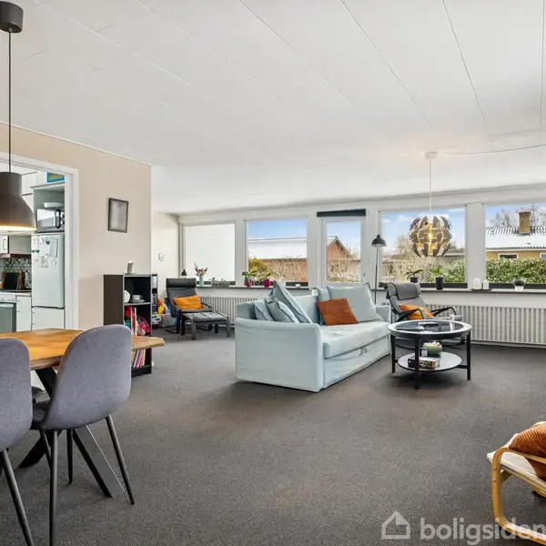 Stue med lyseblå sofa, placeret i et rum med store vinduer, der viser en grøn udsigt. Rundt om sofabordet er stole, og der er også et spisebord med stole.