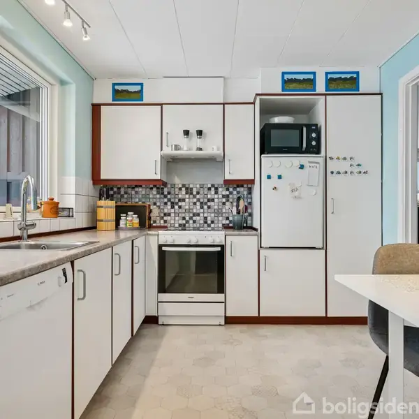 Køkken med hvide skabe og grå bordplade, komfur i midten og mosaikfliser bagved. Mikrobølgeovn ovenpå højre skab. Et bord med stole forrest. Store vinduer giver lysindfald.