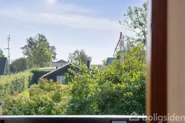 Vindue med udsigt til grøn have og huse. Træer og buske omgiver området. I baggrunden ses en kran og den klare blå himmel.