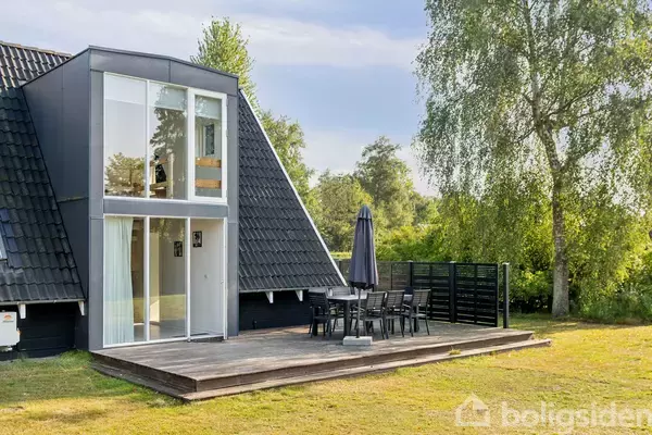Et moderne, sort-hvidt A-formet hus med store vinduer står på en græsplæne. En udendørs terrasse med bord, stole og parasol er placeret ved siden af huset.